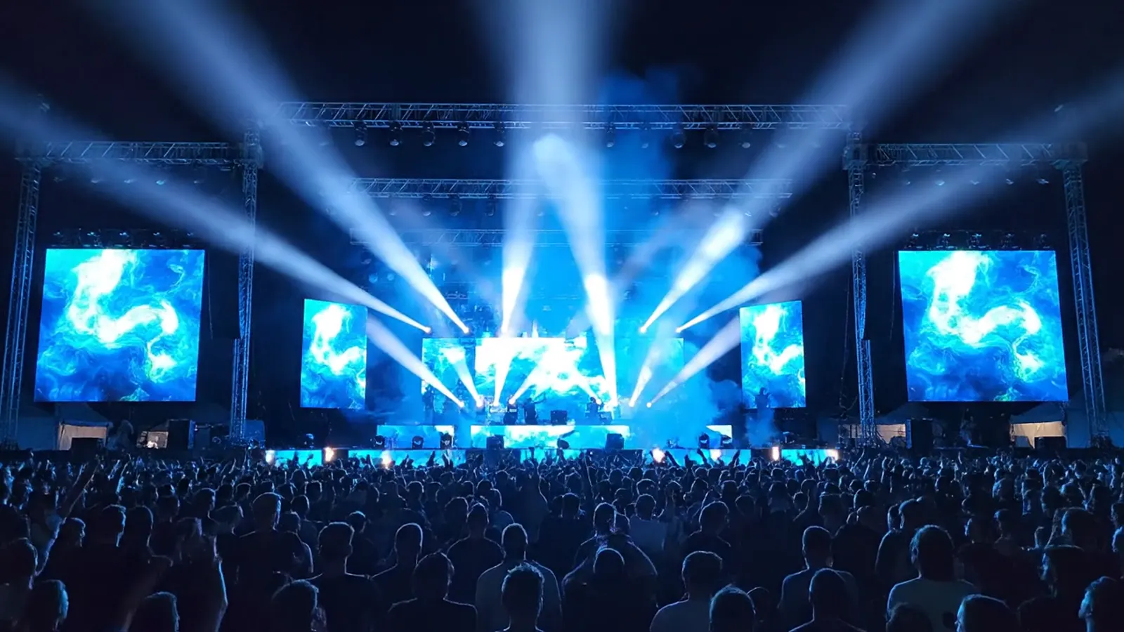 An outdoor stage with truss lights, screens and sparks over a crowd